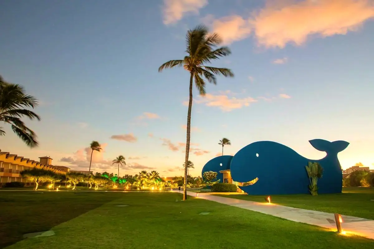 Escultura de baleia azul no gramado ao pôr do sol, entre coqueiros e o resort iluminado
