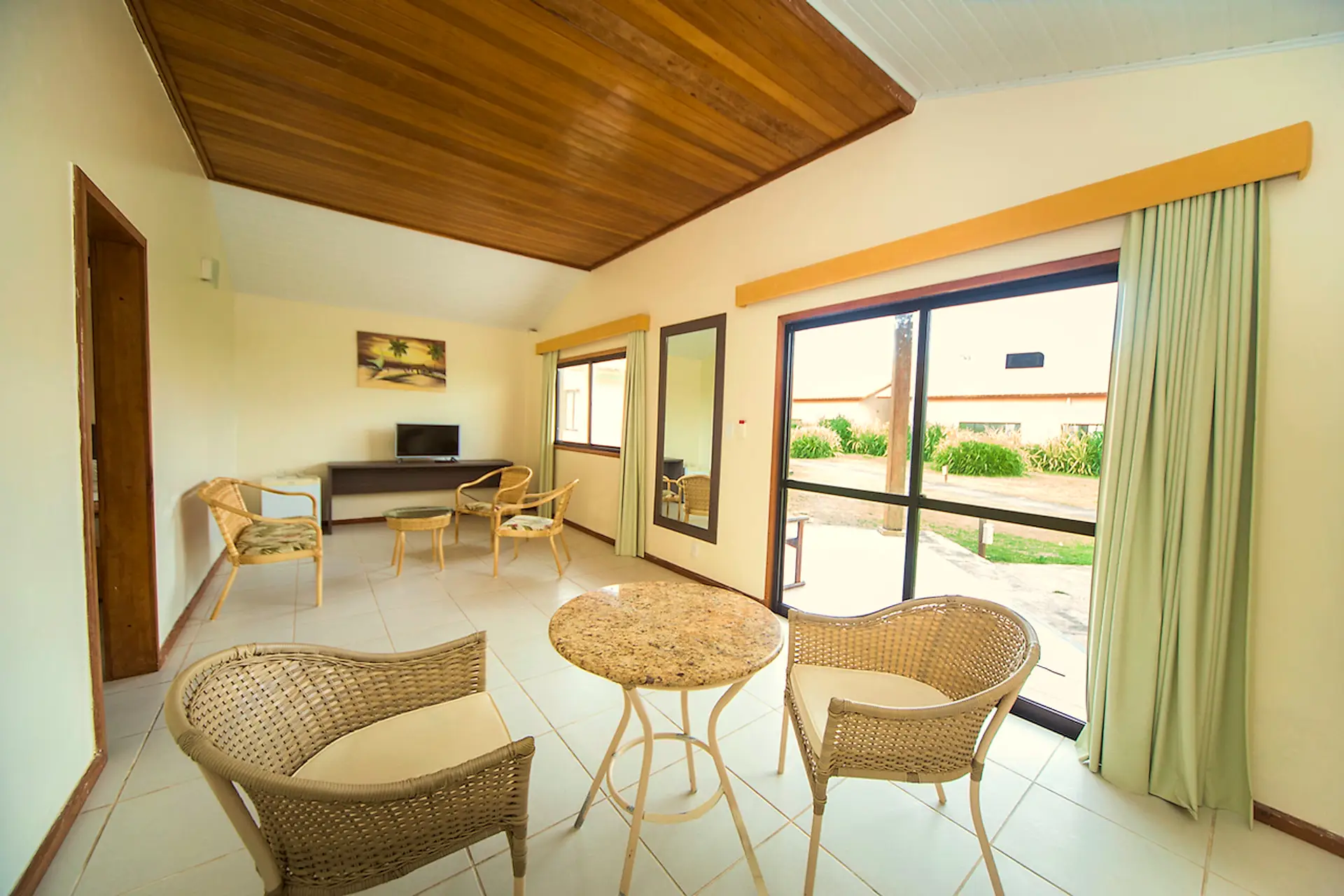 Sala do bangalô com mesa redonda, poltronas de vime e porta de vidro para a varanda