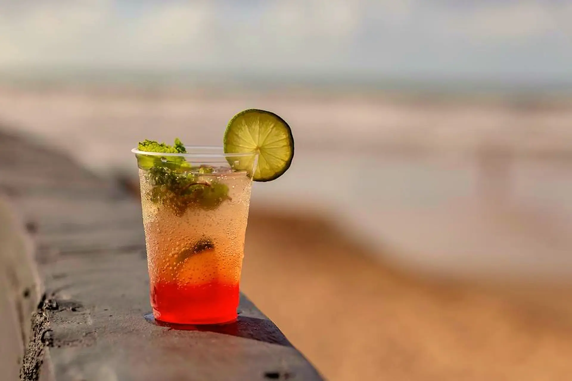 Drink com hortelã, limão e xarope vermelho sobre a mureta, com a praia ao fundo