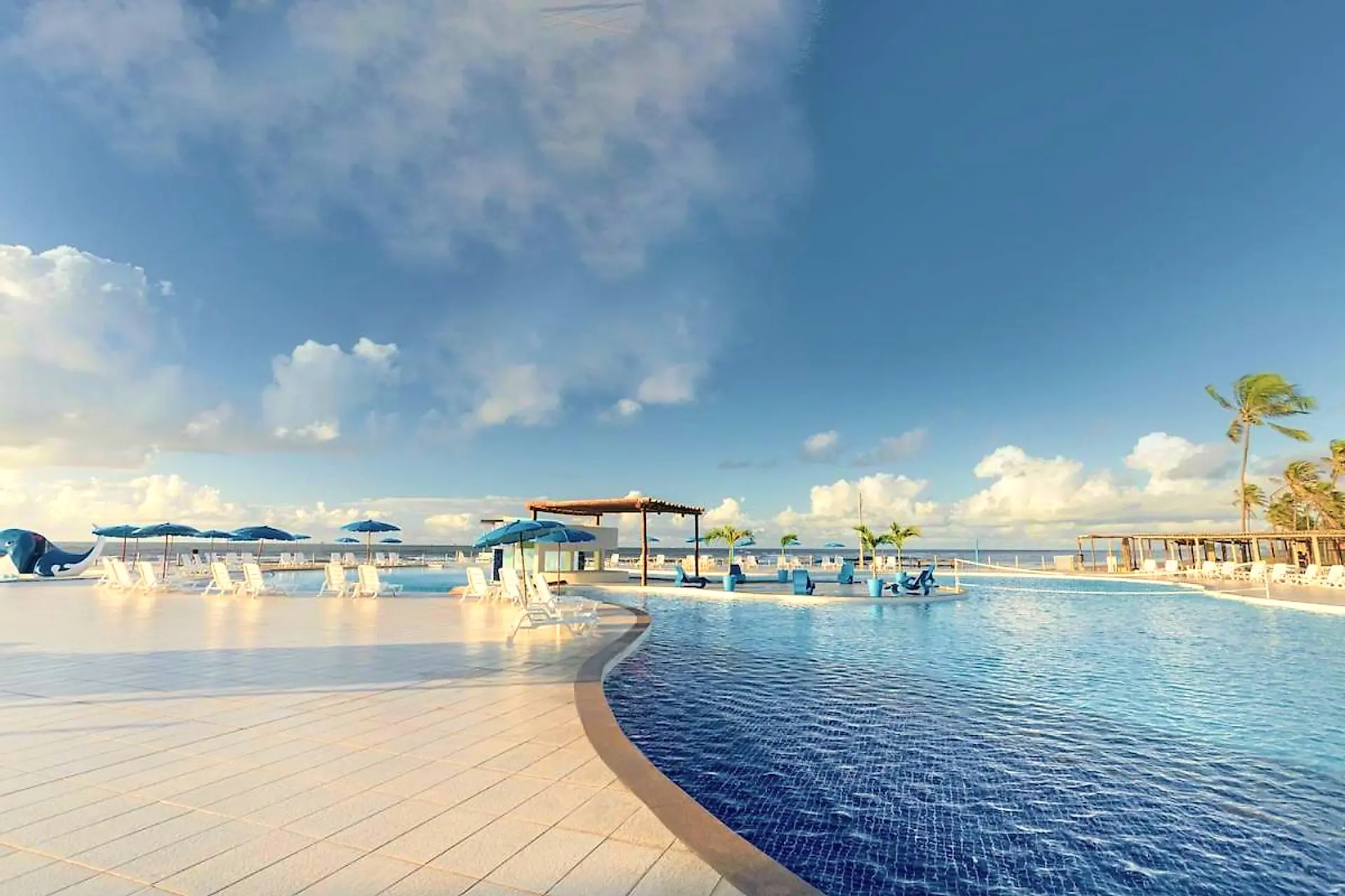 Grande piscina de borda sinuosa com espreguiçadeiras e guarda-sóis azuis sob o céu com nuvens