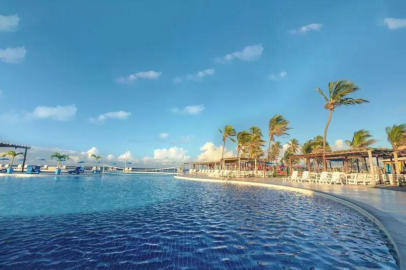 Piscina azul com coqueiros e quiosques de palha ao redor sob o céu aberto