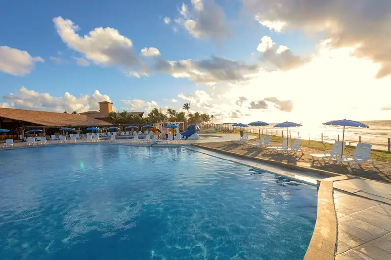 Piscina grande com toboáguas ao fundo, espreguiçadeiras e o mar à direita