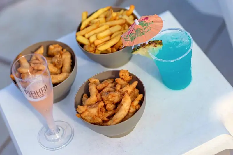 Porções de batata frita e petiscos com drink azul e taça sobre a mesa branca