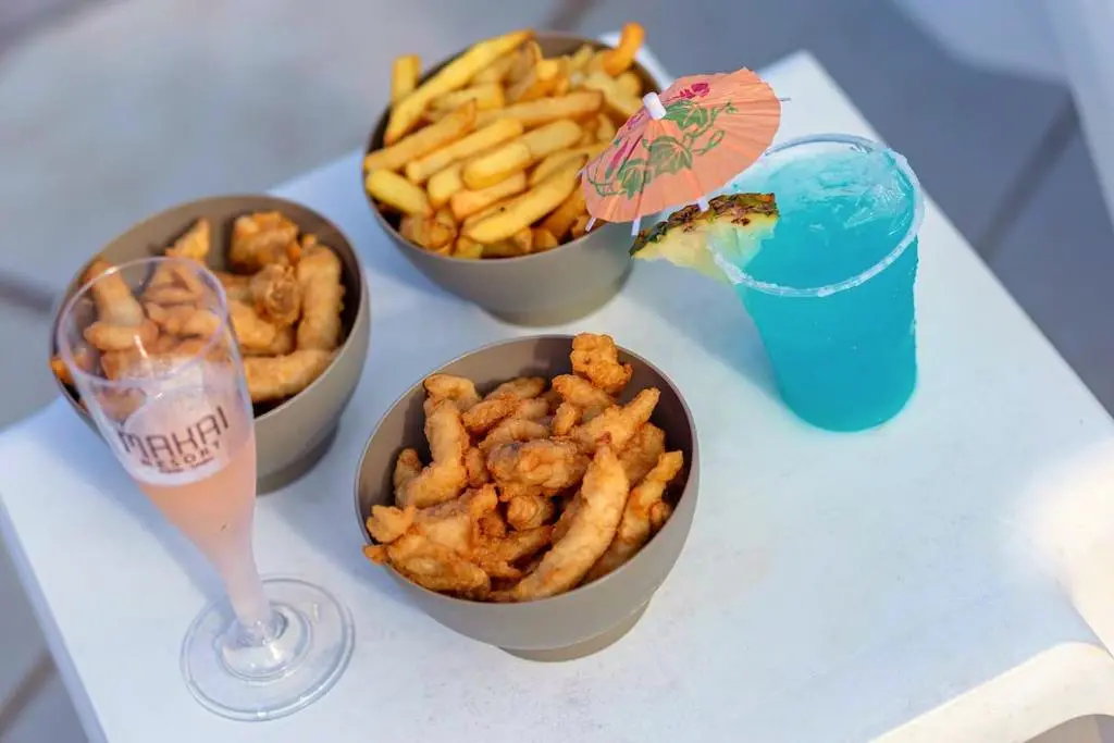 Porções de batata frita e petiscos com drink azul e taça sobre a mesa branca