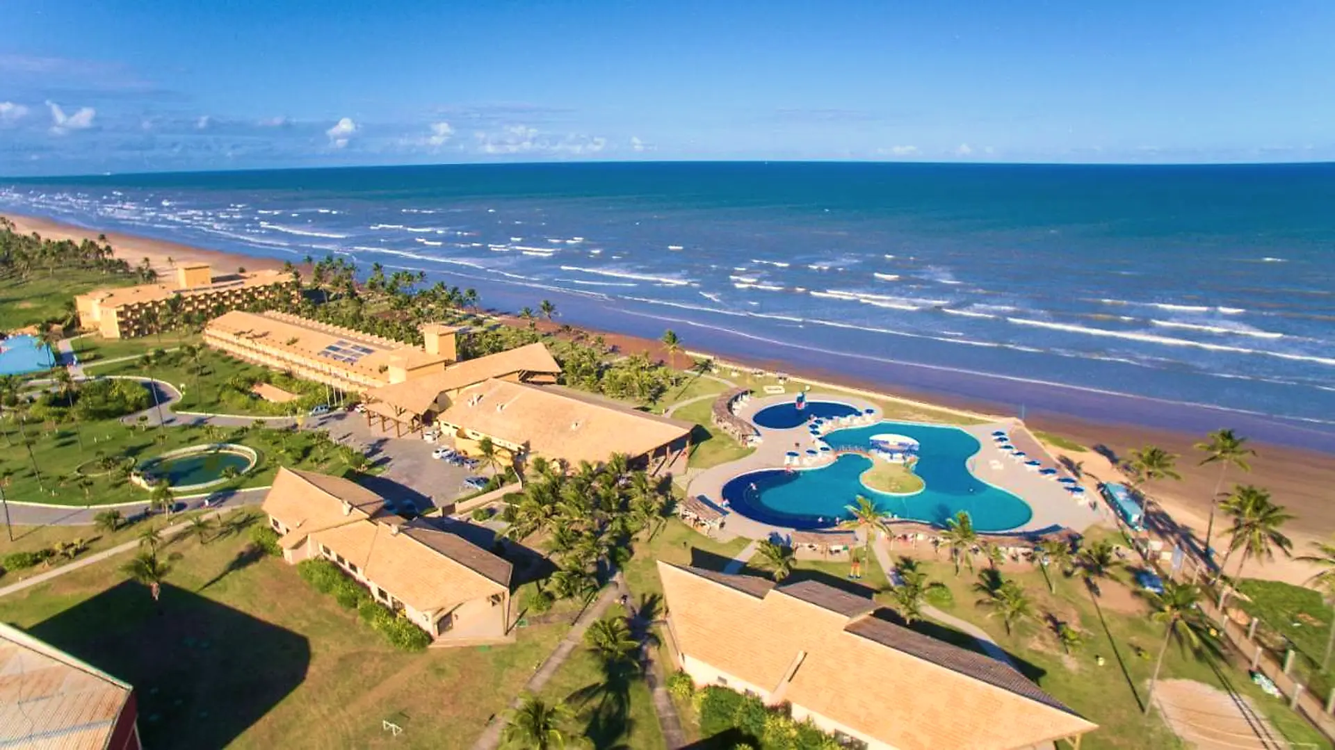 Vista aérea do resort com as piscinas, os blocos de apartamentos e o mar ao fundo