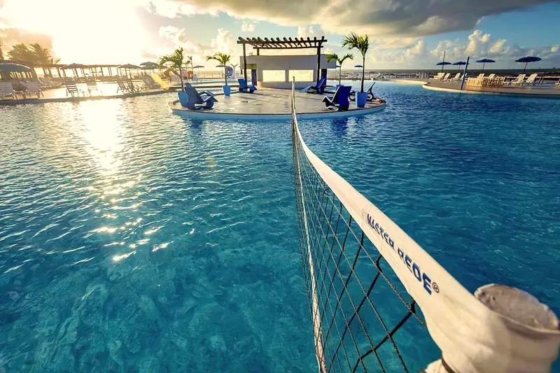 Rede de vôlei aquático sobre a piscina azul, com pergolado e coqueiros ao fundo